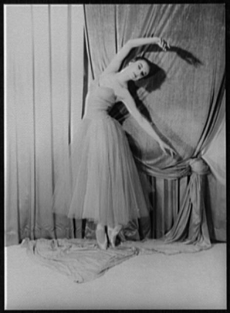 Female ballet dancer in classical tutu holds an extended arabesque, one leg raised behind, arms framing her lifted torso, against draped fabric backdrop.