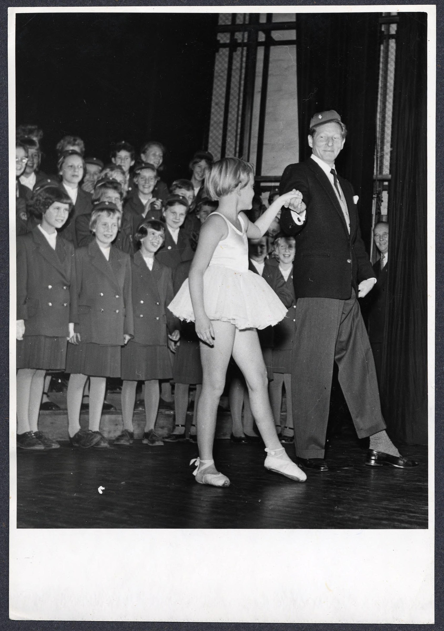 Man in dark suit and fedora holds hands with young woman in white dress on stage before an audience of formally dressed women.