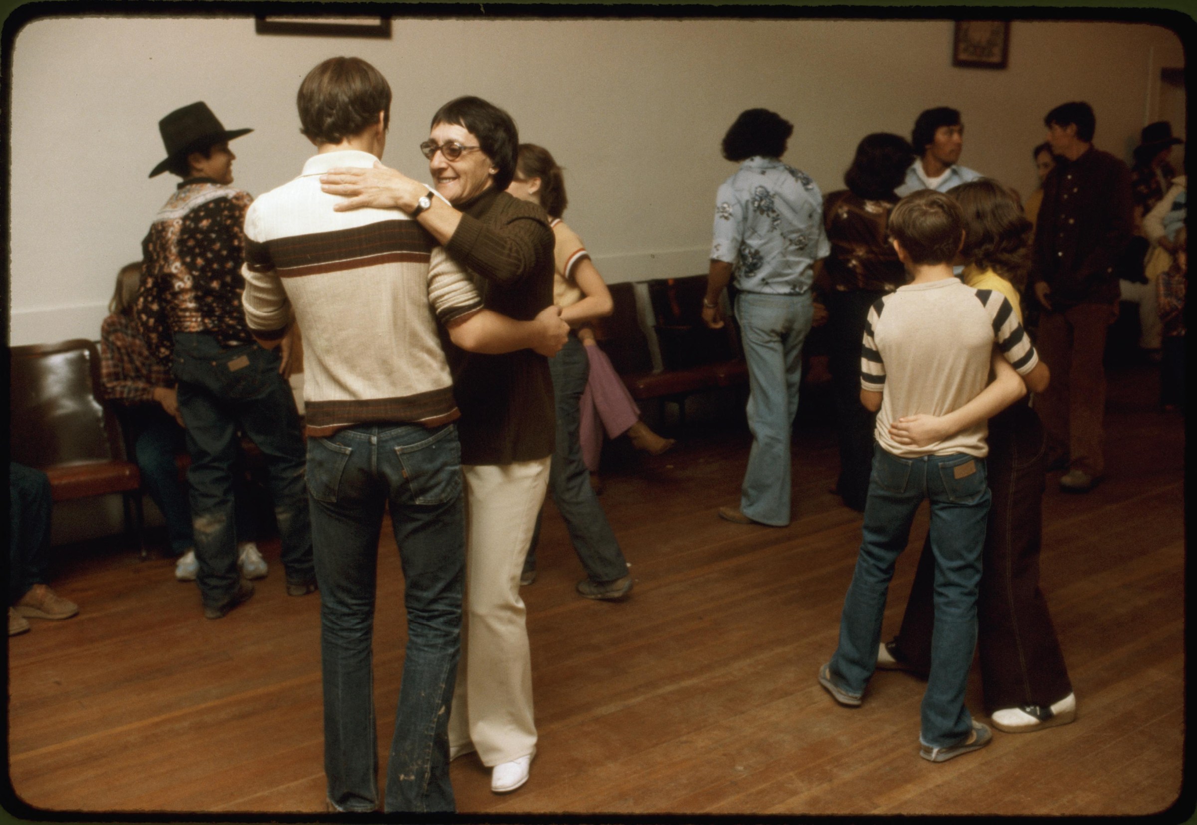 A woman in glasses and brown cardigan dances with a young boy in a striped shirt in a wood-floored community hall, with other children in pairs around them.