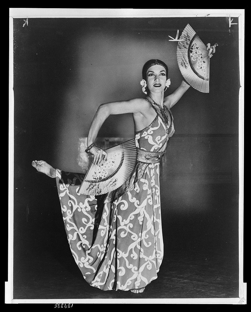 Female performer in decorative patterned costume and pearl earrings holds an ornate pleated fan, posed on a dark stage in 1944.