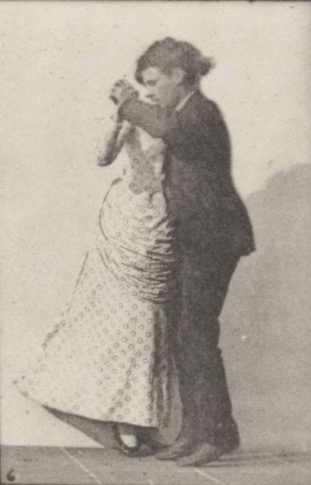 A man and woman in Victorian dress perform a waltz in closed ballroom position, captured mid-turn from the side.