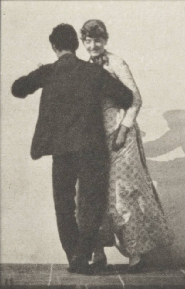 Man and woman in formal dress waltzing together in closed ballroom hold, photographed from the front against a plain background.