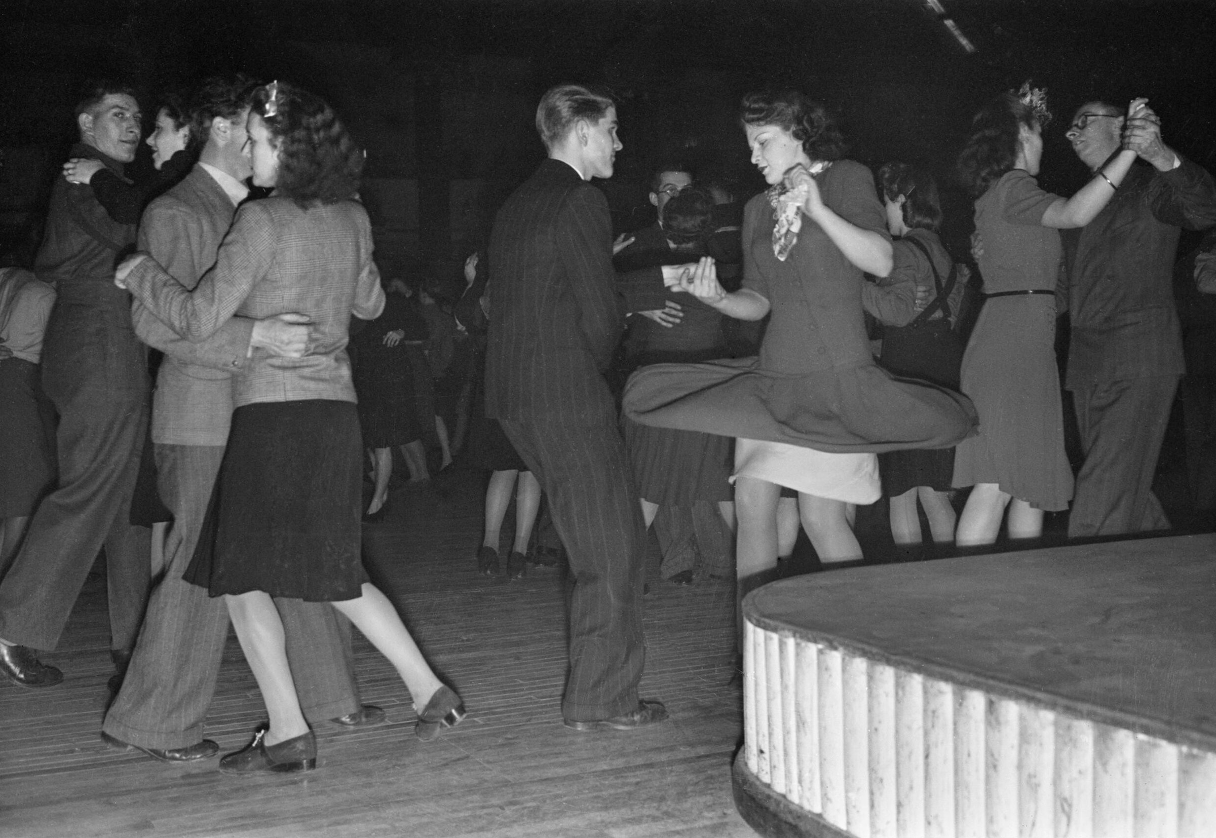 Black-and-white photograph of a 1945 British dance hall: a couple in the centre executes dynamic jive with swinging legs and loose hold, surrounded by formally