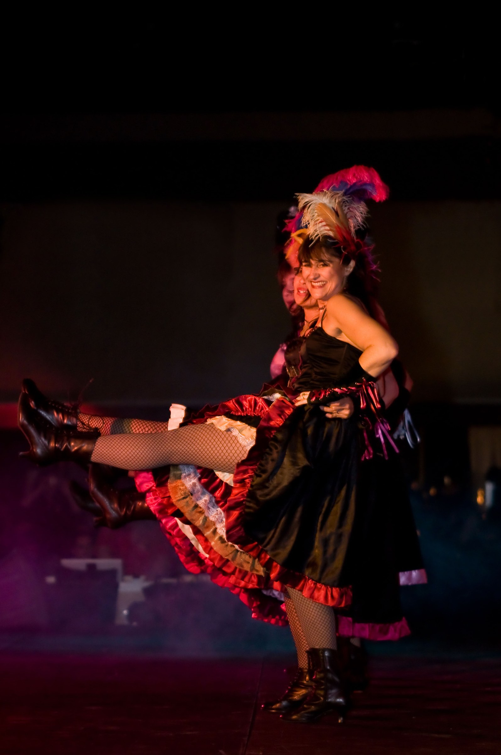 A female dancer in a black corset and layered red ruffled skirt performs a high kick, smiling at the camera, wearing a tall feathered headdress under warm