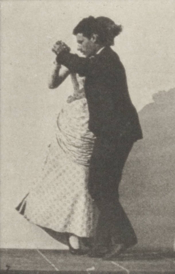 Man in dark formal dress and woman in light pleated dress demonstrate ballroom waltz hold and posture, photographed against plain backdrop.
