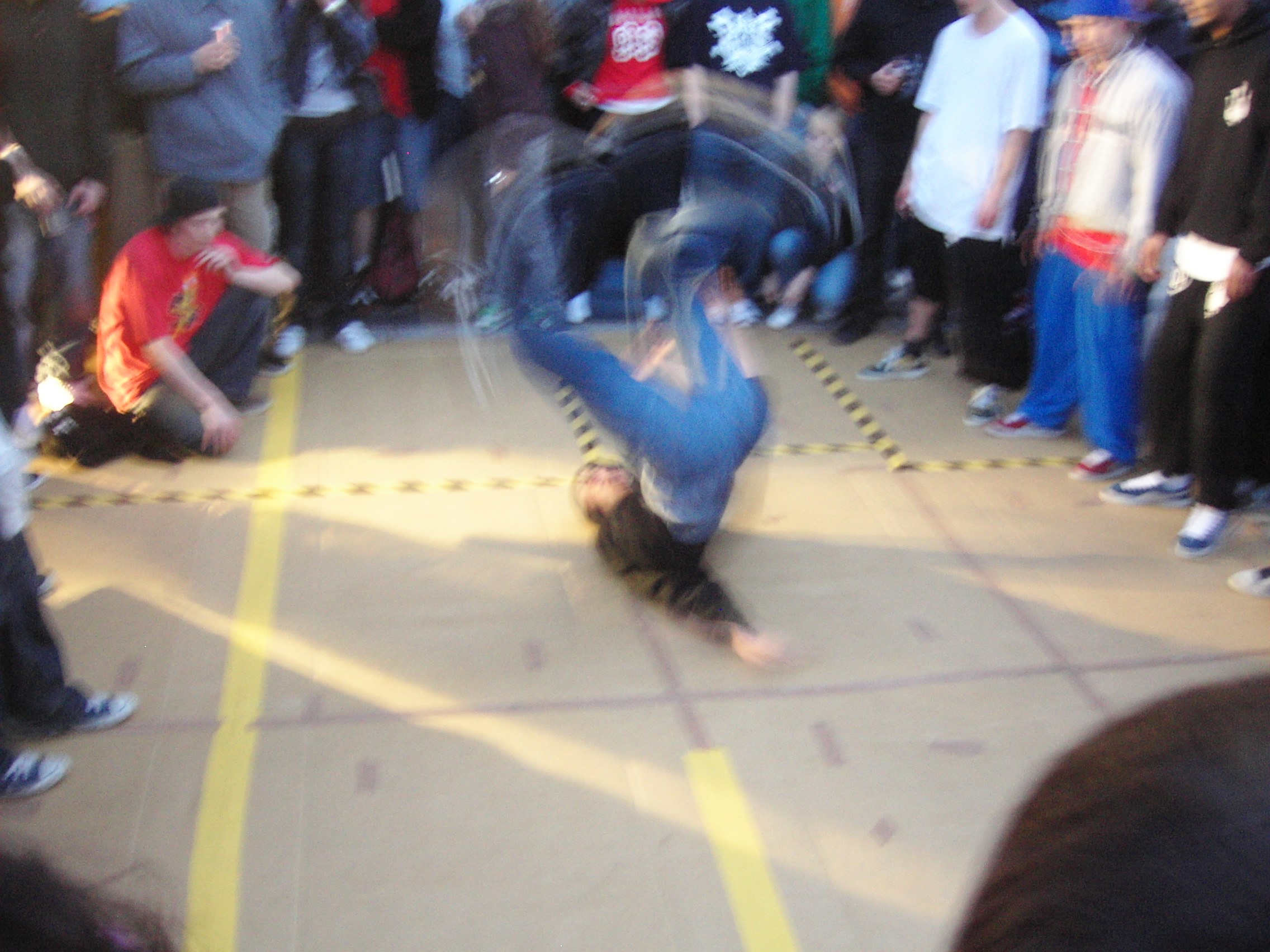 Breakdancer inverted in a freeze move within a yellow-taped circle, surrounded by standing spectators on a city street.