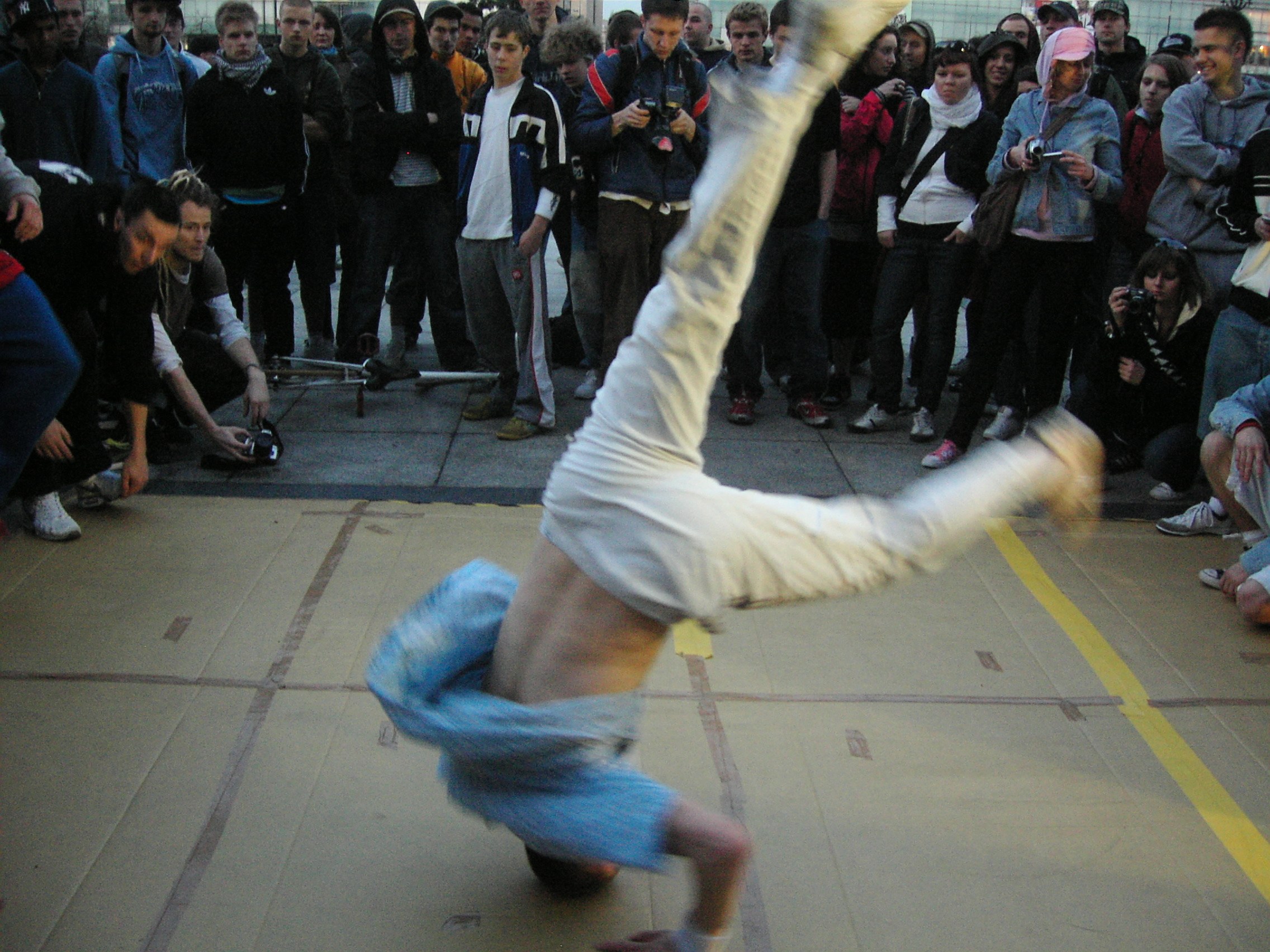 Breakdancer performs inverted power move in gymnasium surrounded by youth crowd holding cameras.