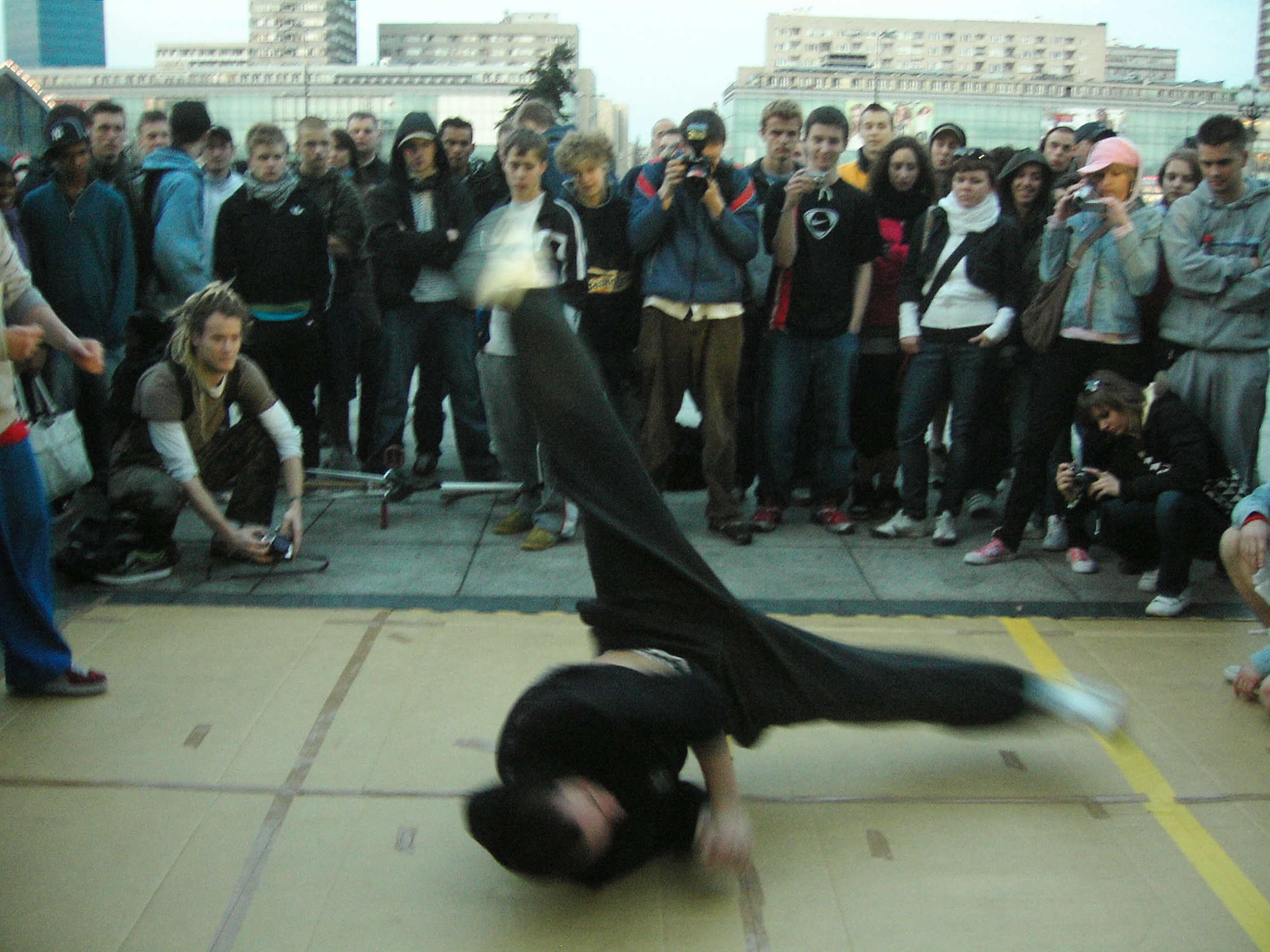 A breakdancer performs an inverted freeze on concrete while a crowd of young people records on mobile phones in an urban plaza.