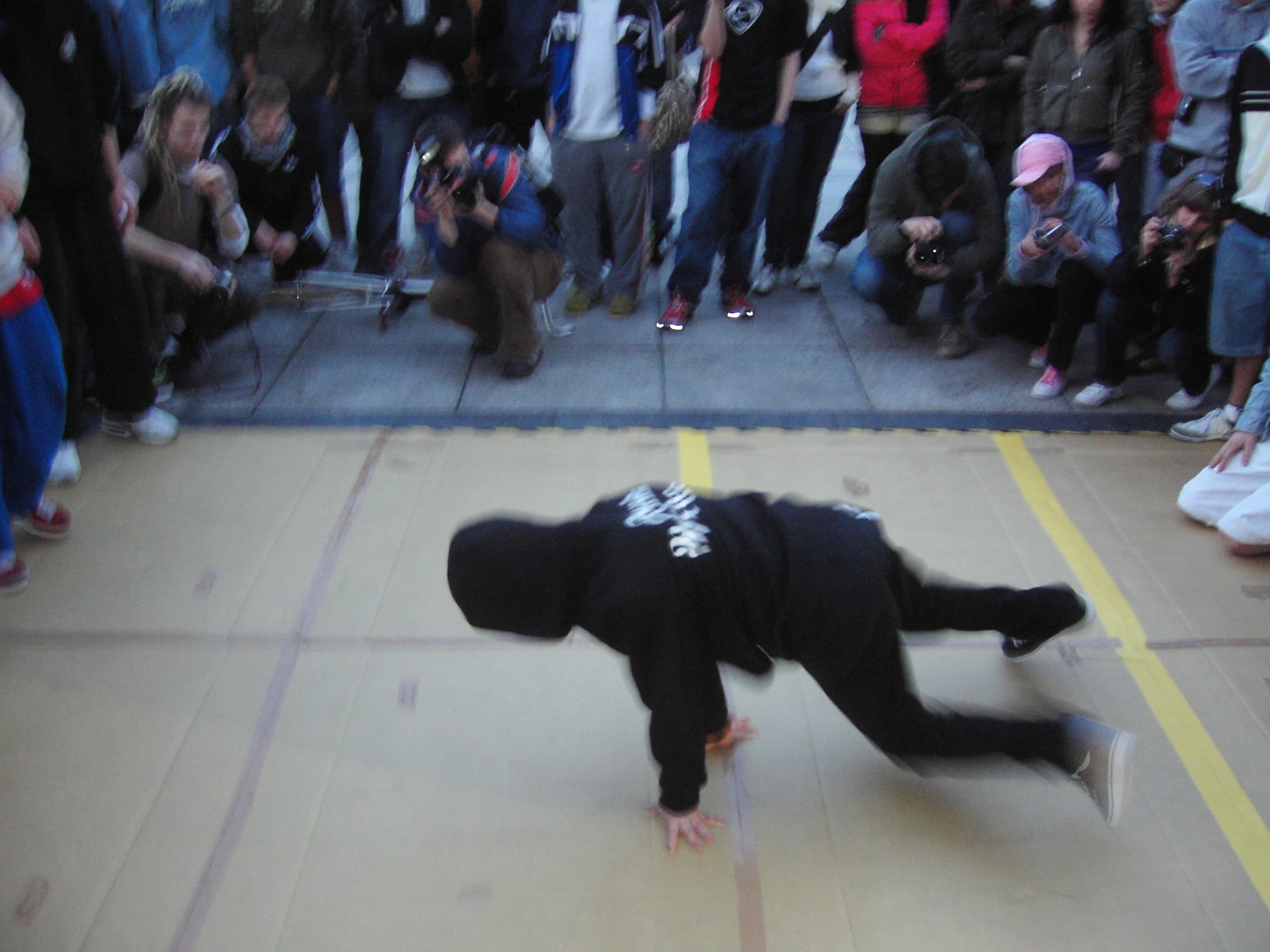 A male breakdancer suspended in an inverted headstand freeze on a gymnasium floor, surrounded by spectators with cameras.