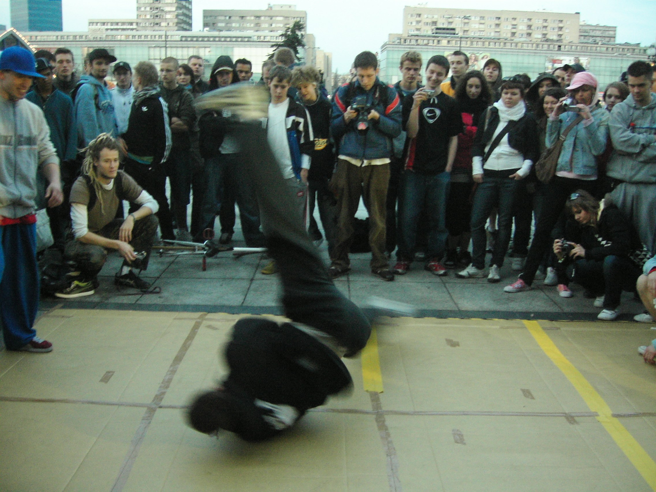 Young breakdancer inverted mid-move in a circle of spectators in a Warsaw plaza, 2007.
