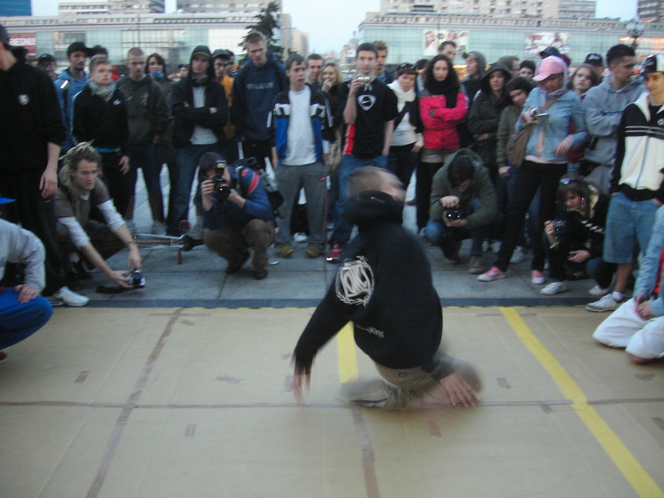 A breakdancer performs an inverted freeze in the center of a public plaza, surrounded by spectators holding phones.