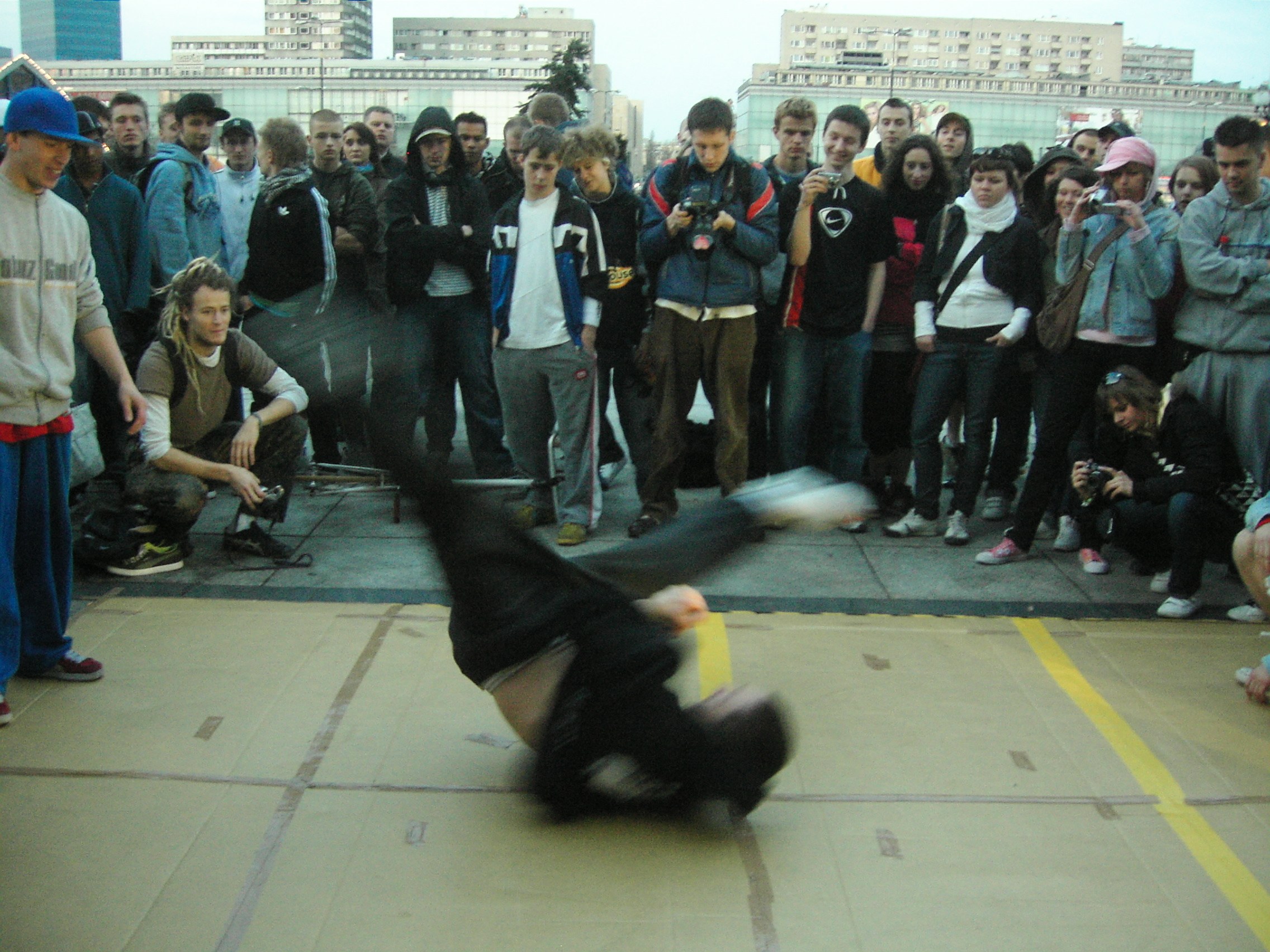 Breakdancer spinning inverted in center of concrete plaza surrounded by watching crowd; Soviet apartment blocks in background