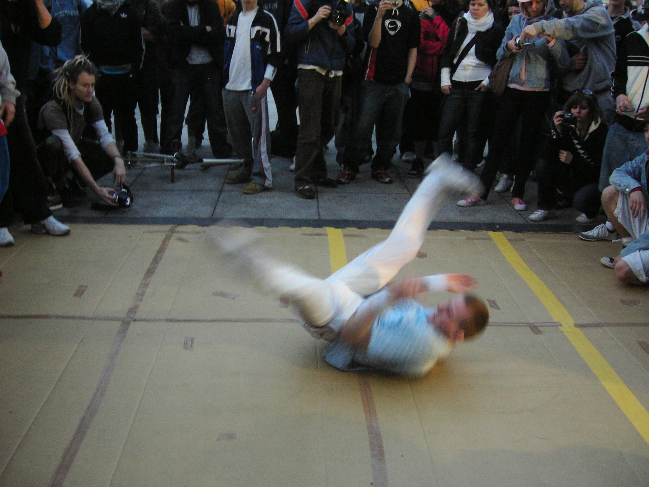 Breakdancer performing an inverted freeze move on a gymnasium floor surrounded by spectators holding cameras.