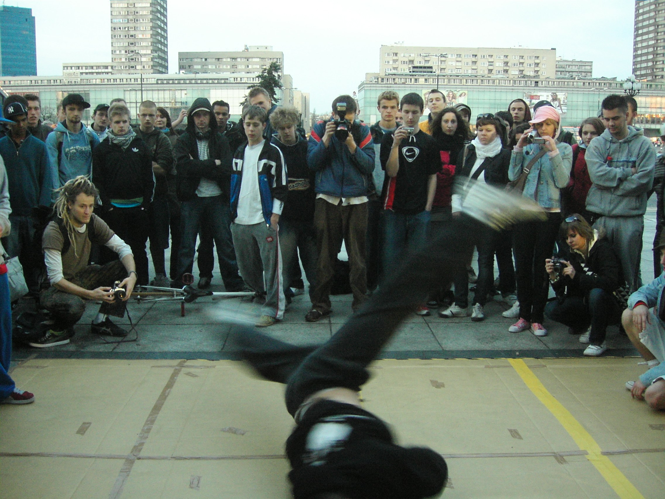 Breakdancer performing an inverted freeze in a concrete plaza, surrounded by a crowd of young spectators holding cameras.