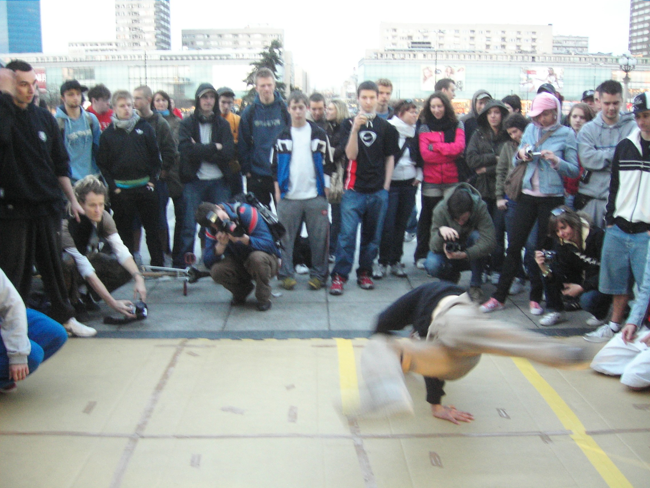 Two breakdancers perform on a Warsaw street: one horizontal in a freeze, the other crouching nearby, surrounded by spectators with mobile phones.