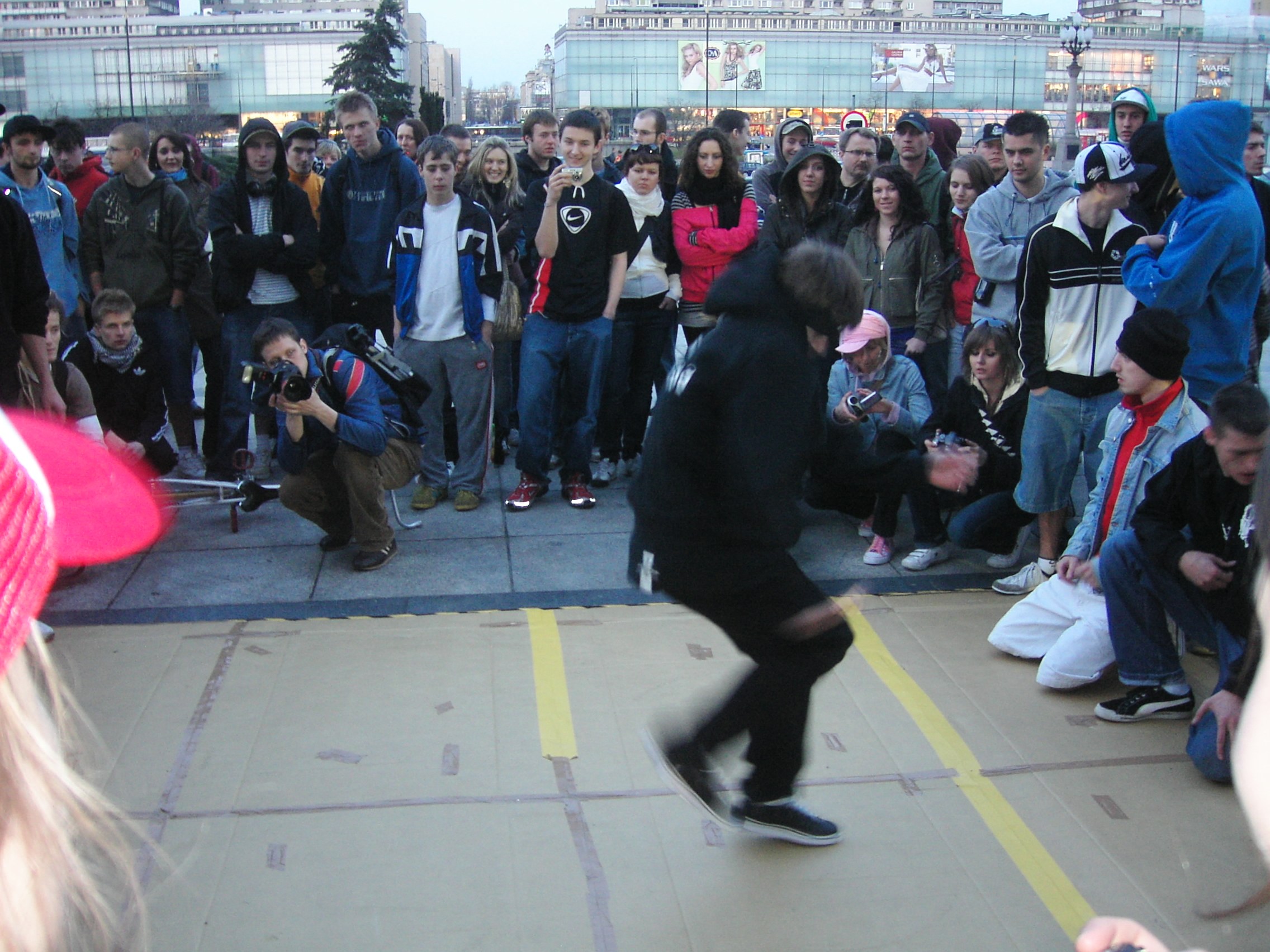 A dancer in black performs breaking in a paved urban plaza while roughly 40 spectators stand in a circle, many recording on mobile phones.