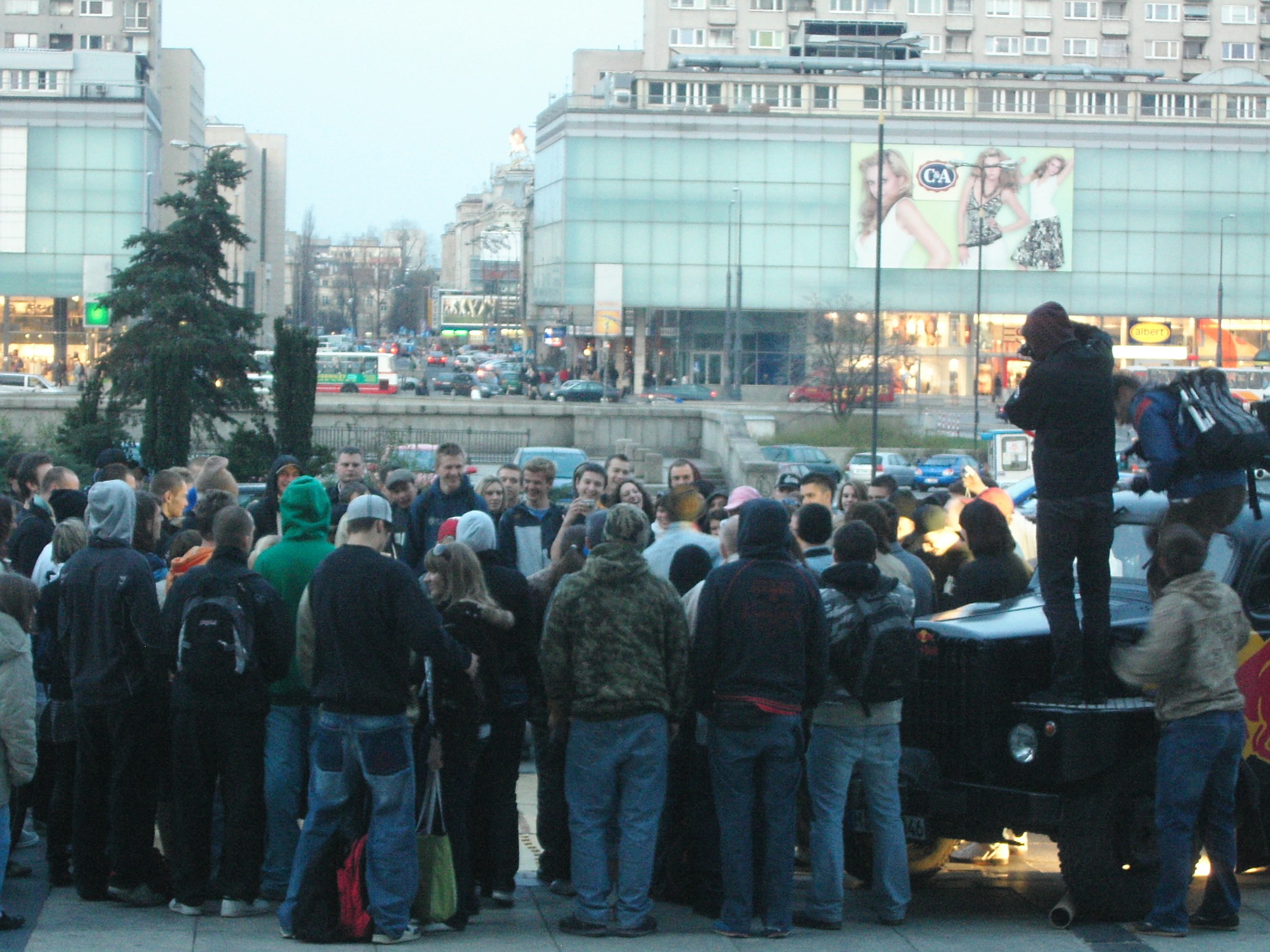 Crowd of young people in winter clothing gathered in Warsaw plaza around a breakdancing cypher, with residential buildings and C&A store visible behind them.
