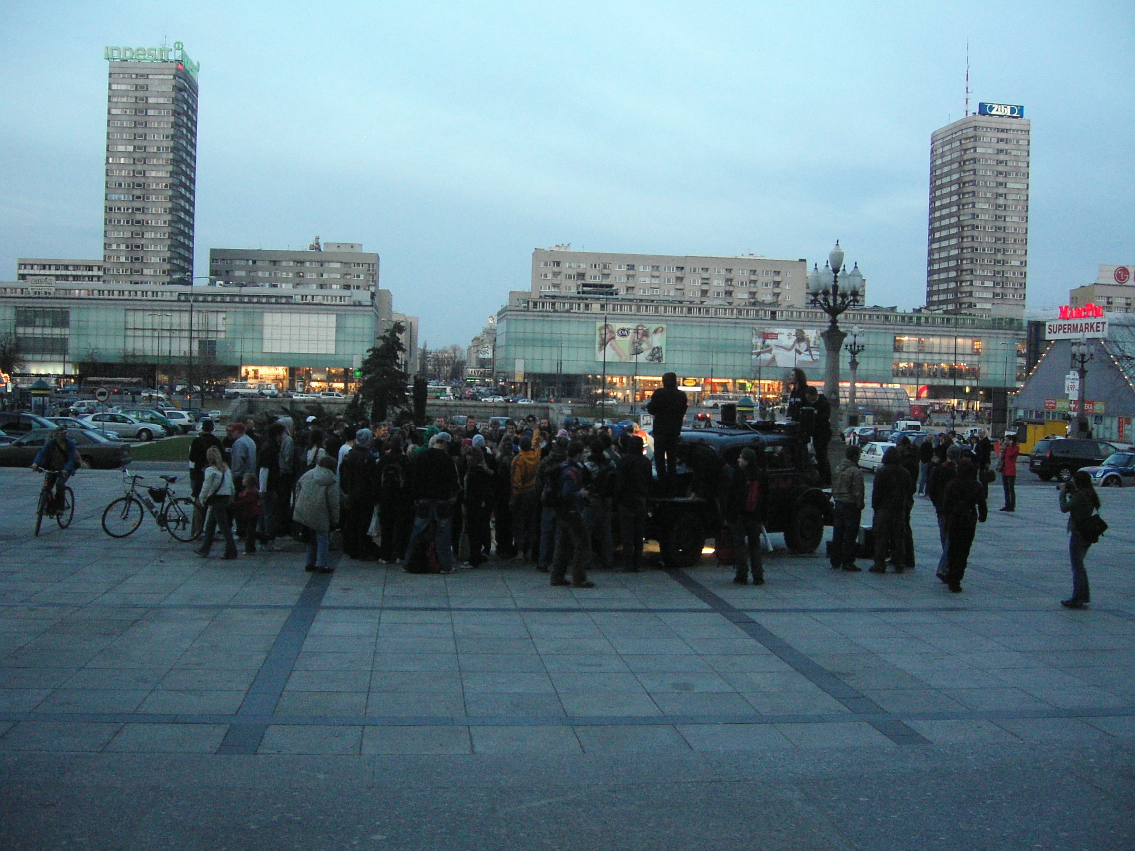Dense crowd gathered in a lit urban plaza at dusk, with figures in dark winter clothing clustered around a central cleared space.