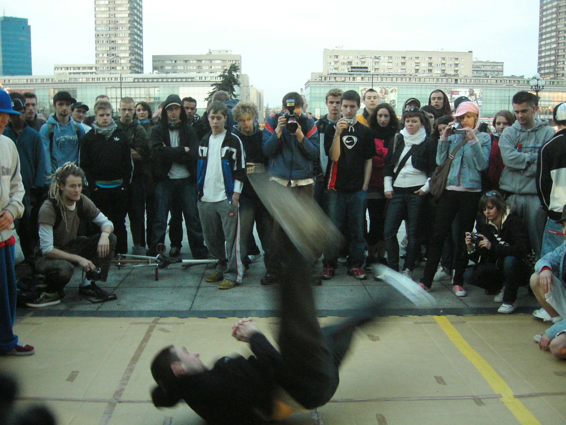B-boy performs an inverted freeze in a Warsaw street cipher surrounded by spectators recording with cameras, 2007.