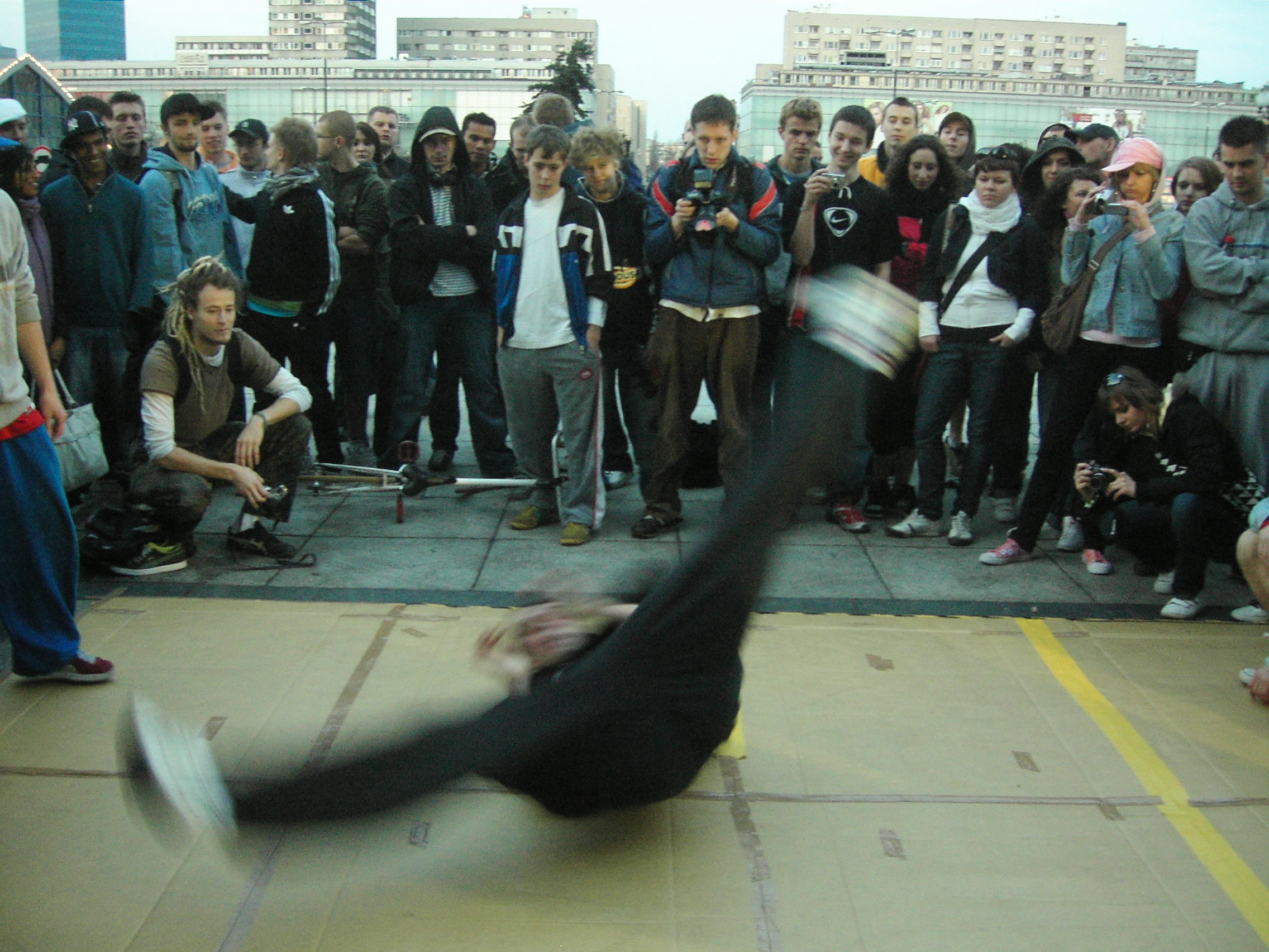 A dancer in horizontal motion blur performs an acrobatic move at the center of a crowd standing in a circle on a concrete plaza.