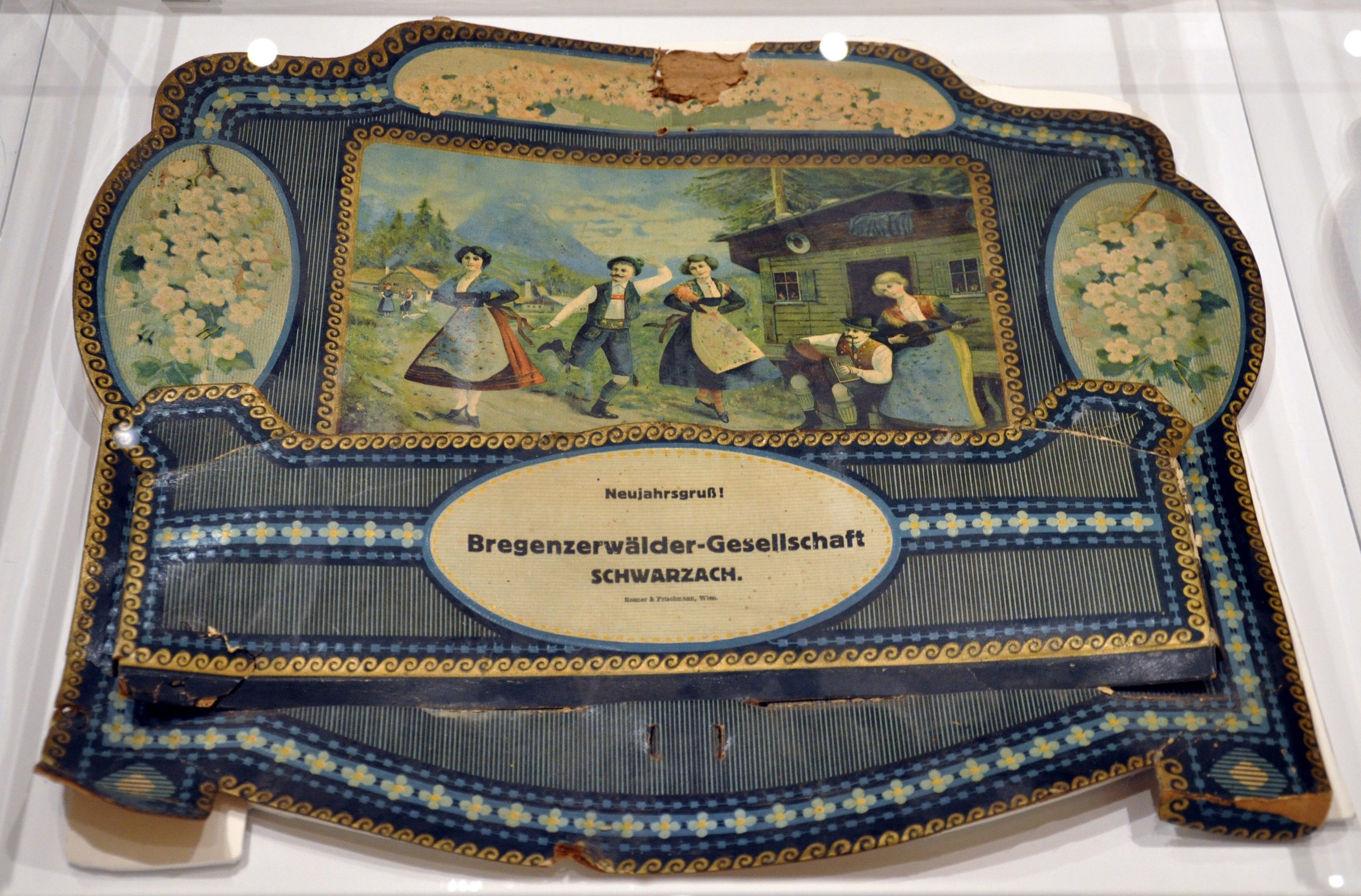 Five people in traditional Austrian Alpine costume dance and celebrate in a rustic cottage, framed by gold and blue decorative borders.