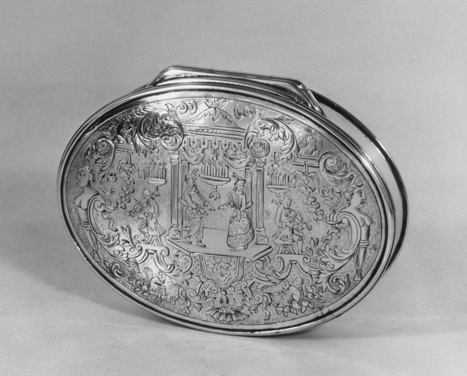 Oval silver snuffbox with relief scene of elegantly dressed figures gathered in a classical interior surrounded by ornate rococo foliage