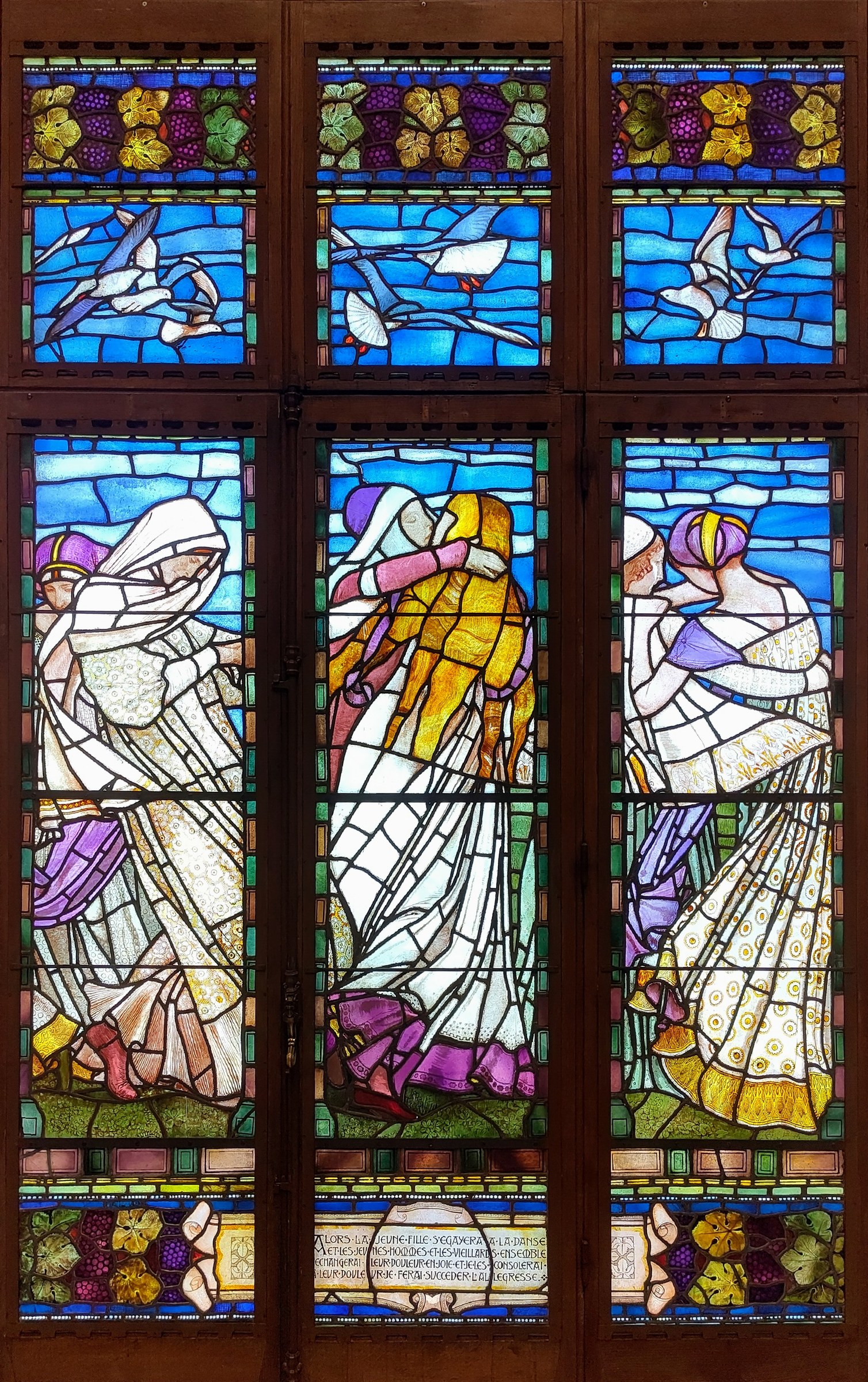 Three tall stained-glass panels depicting flowing female figures in classical drapery against blue backgrounds, separated by lead came.