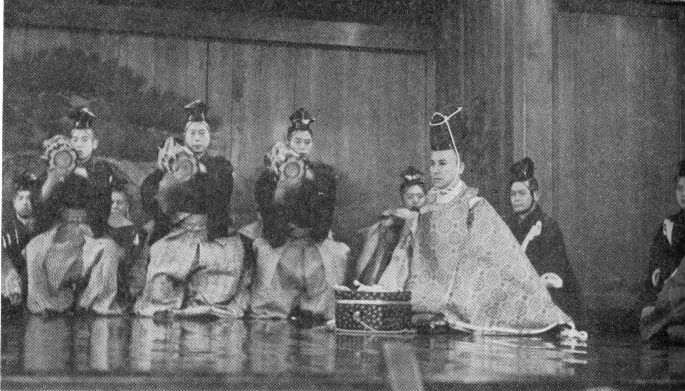 Black-and-white photograph of five Noh theatre performers in formal costumes and masks, arranged in a frontal composition on a bare stage.