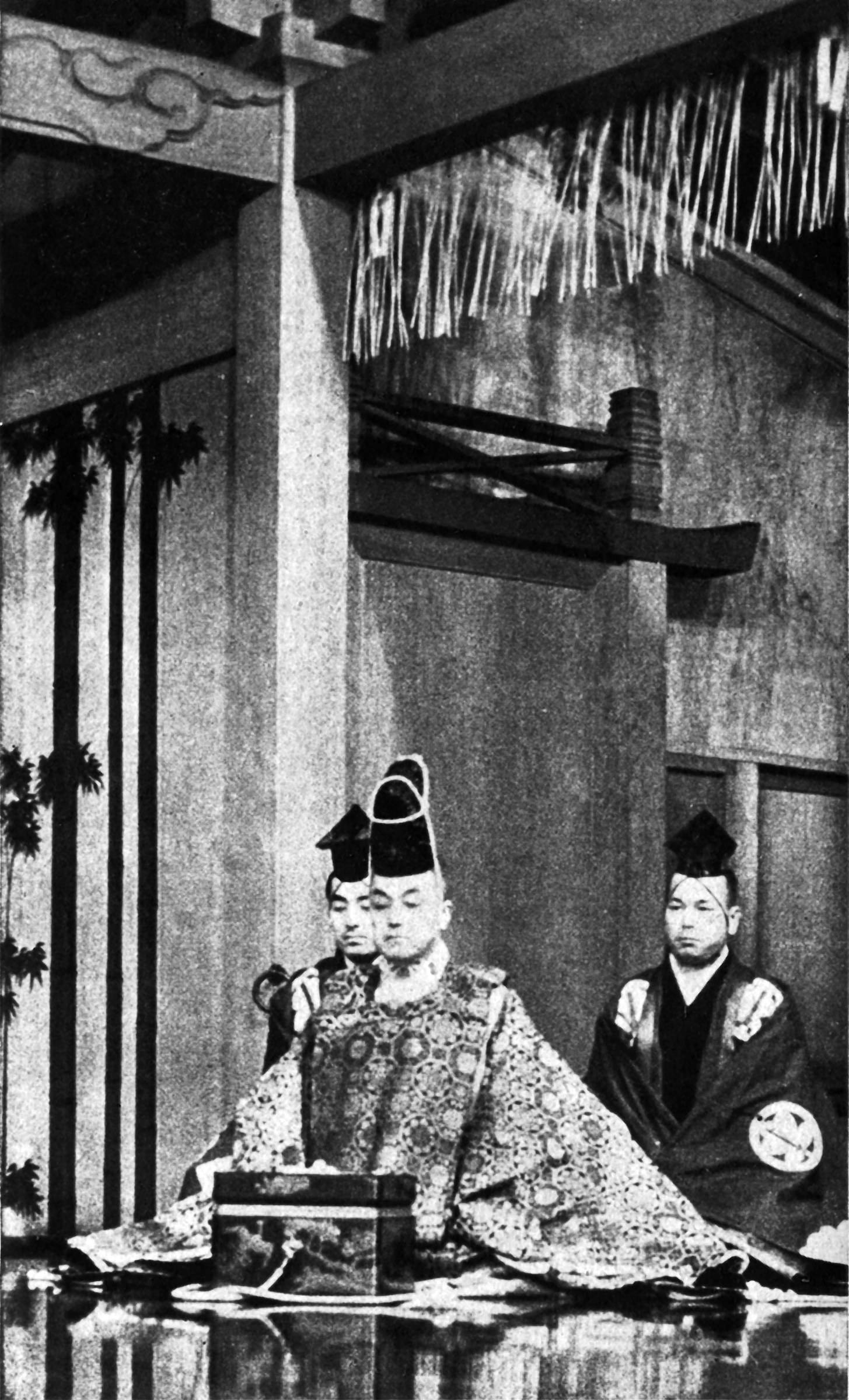 Three figures in formal Noh costume and staging; central figure in brocade robe seated in seiza, wearing tall crown headpiece; attendants flanking; hanging