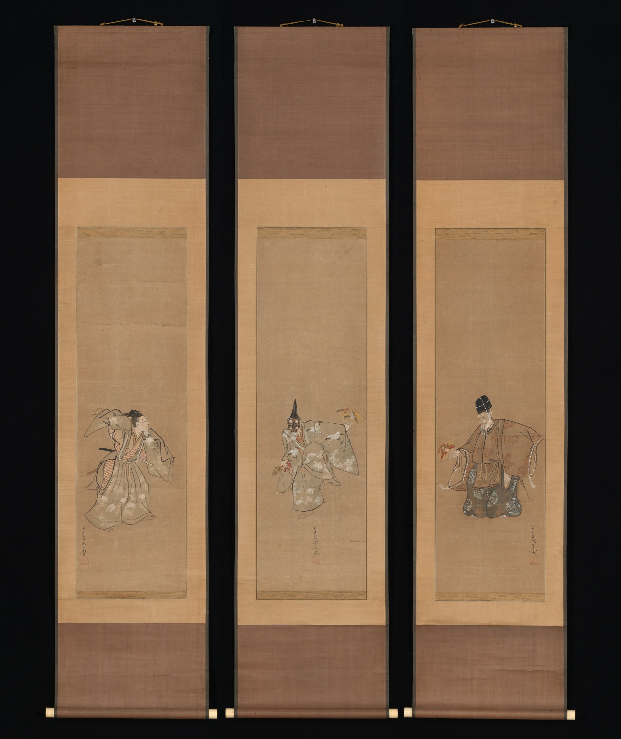 Three Japanese hanging scrolls showing figures in elaborate kimonos and caps, each standing in formal pose with raised arms, representing the Okina Noh dance.