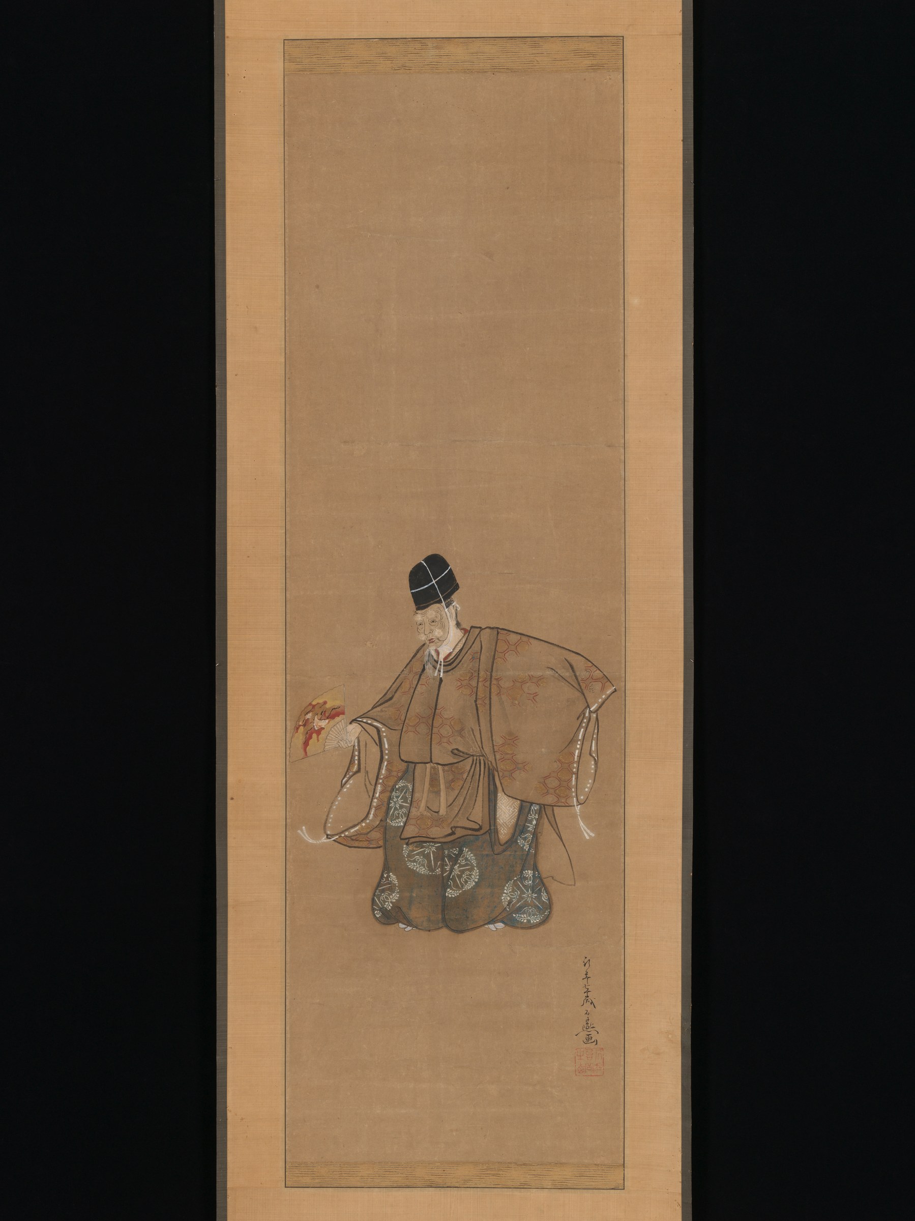 An elderly Japanese man in formal dark robes and patterned garments stands upright, holding a fan, against a plain tan background.