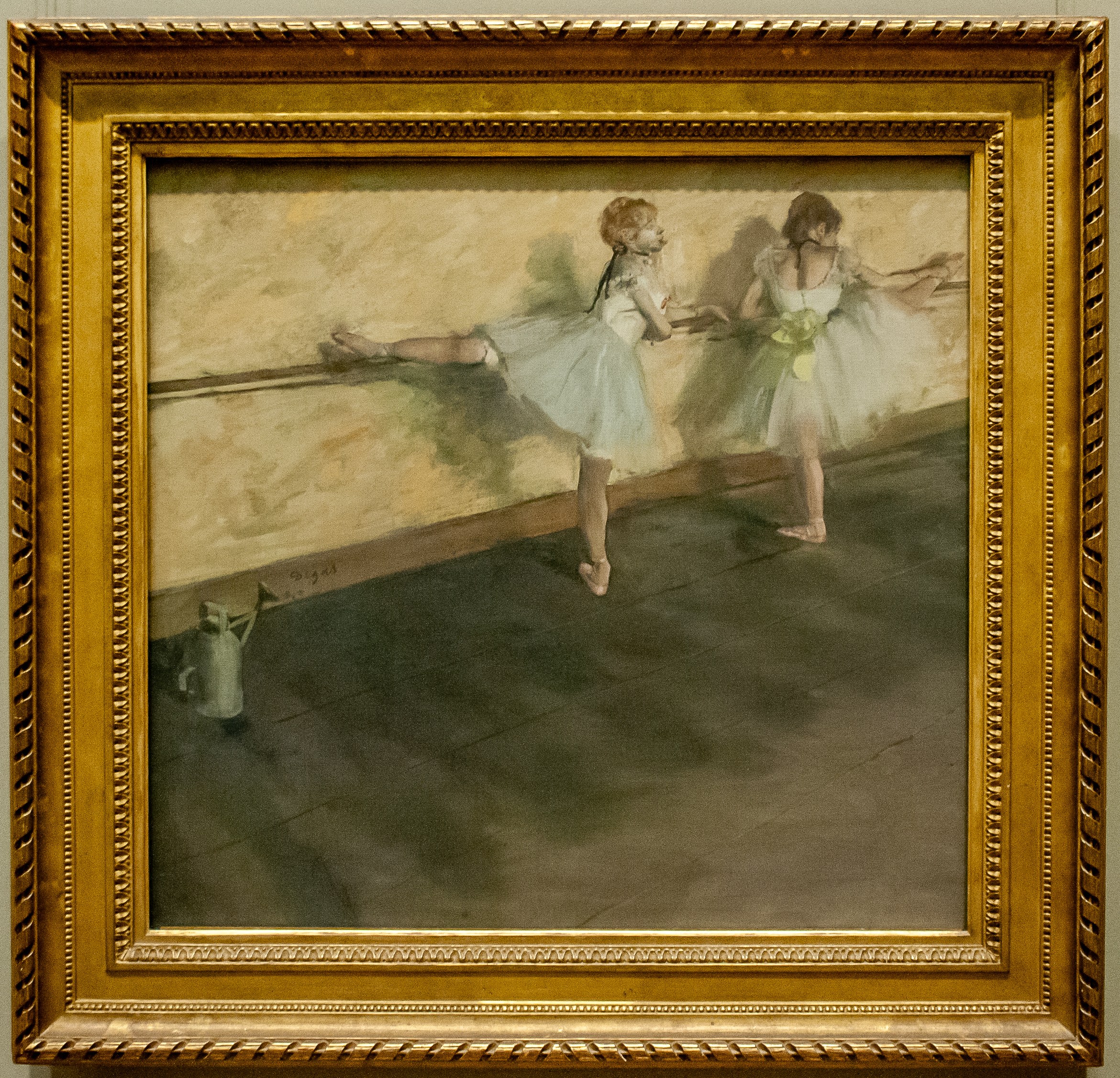 Two female ballet dancers in a studio: one extends her leg while the other guides it, both in pointe shoes and pale practice skirts.