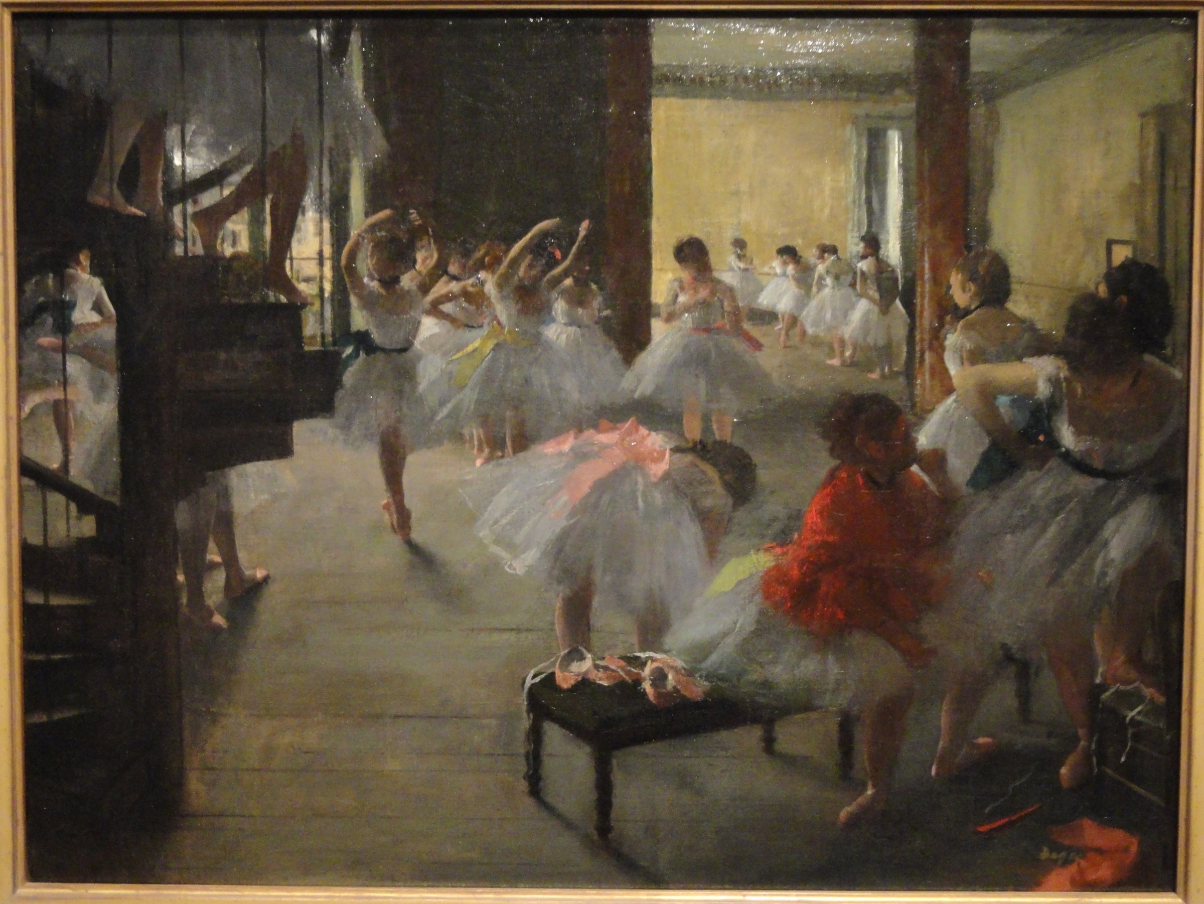 A ballet teacher in dark clothes instructs young female students in white tutus in a sunlit studio, some standing at the barre, one seated on a bench.