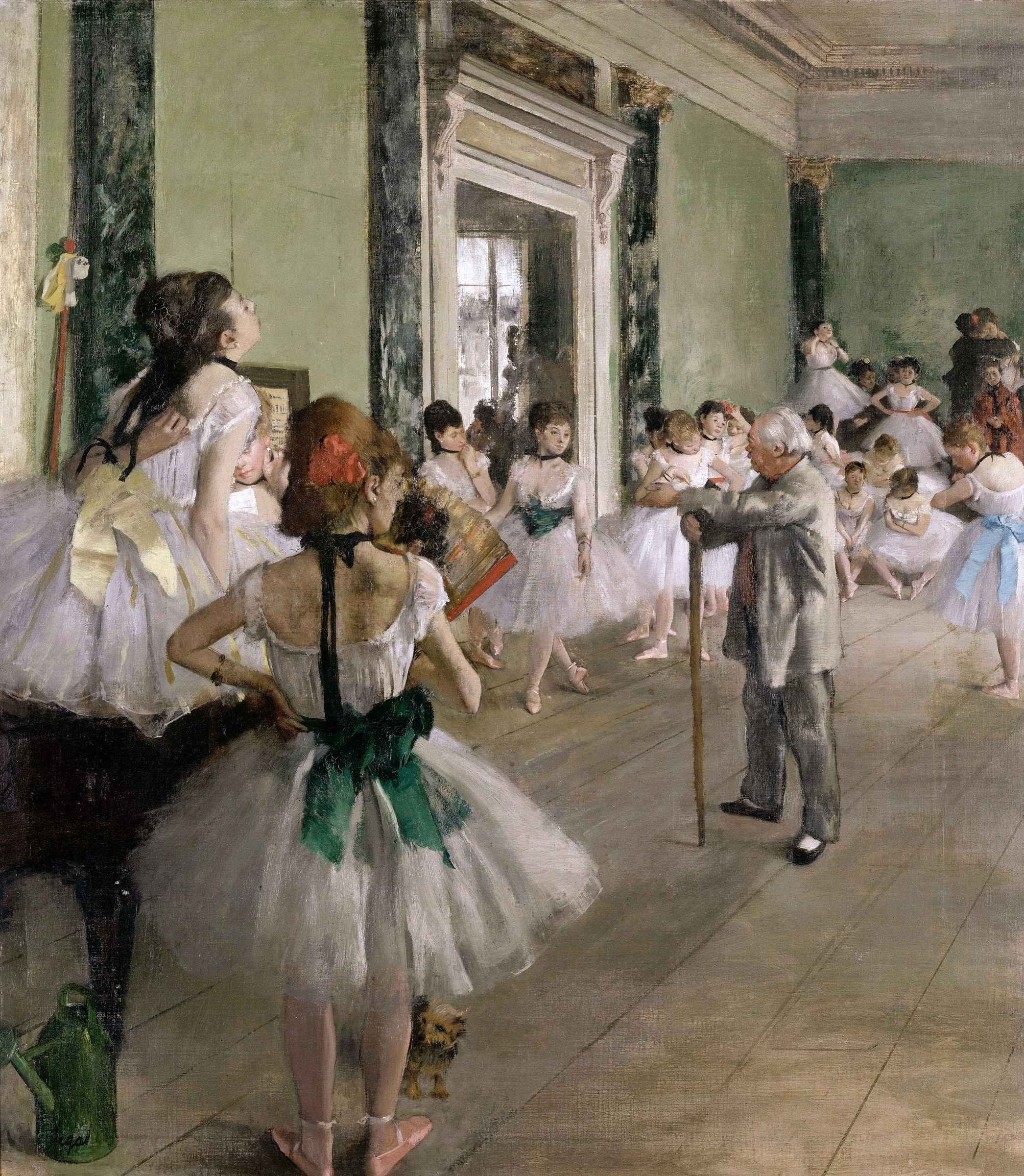 An elderly ballet master in dark coat gestures with a cane to direct a class of about fifteen young female dancers in pale tutus and pointe shoes in a