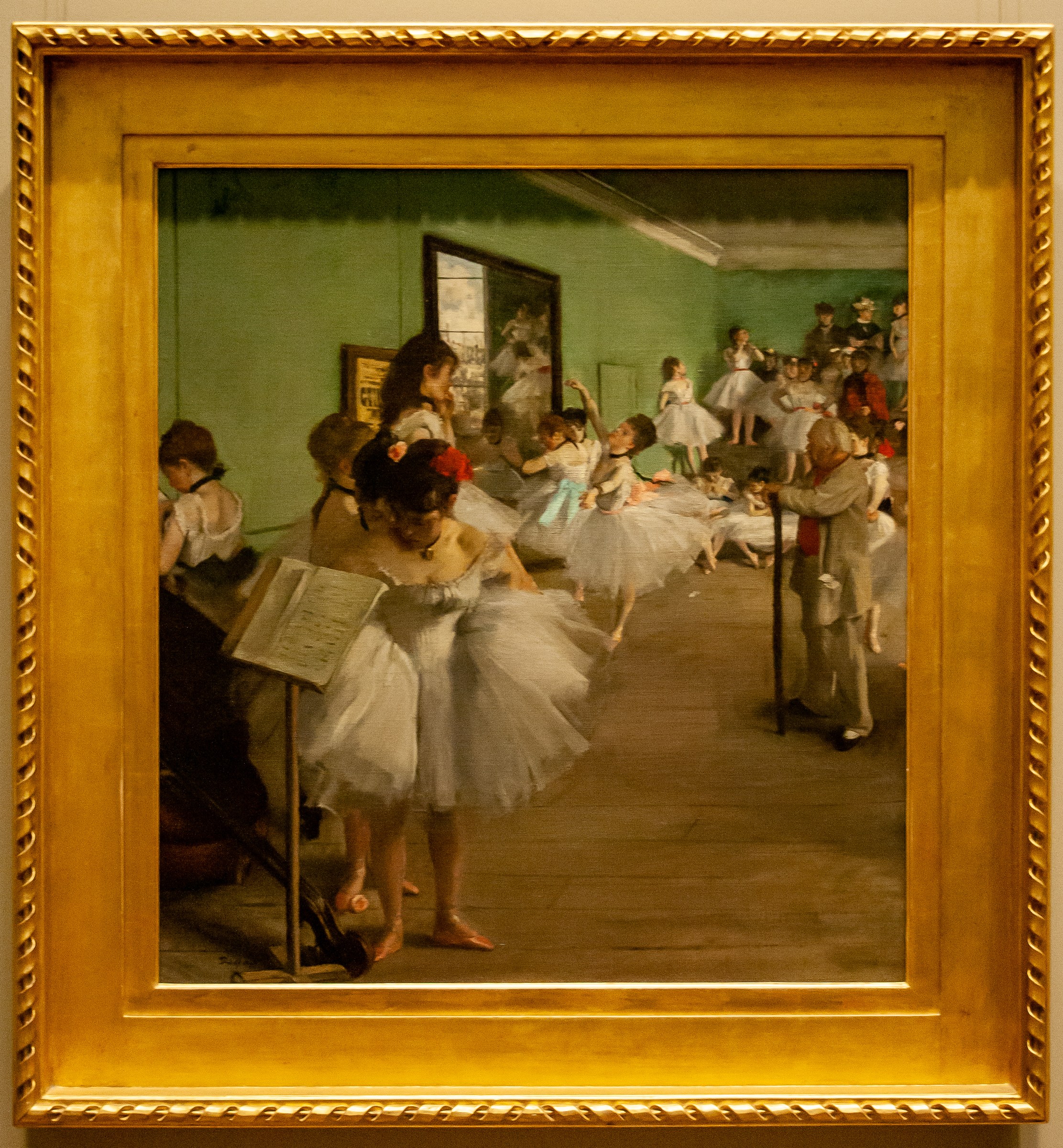 A ballet instructor in dark clothes observes a young dancer in white in a sunlit studio as other students wait or stand in the background.