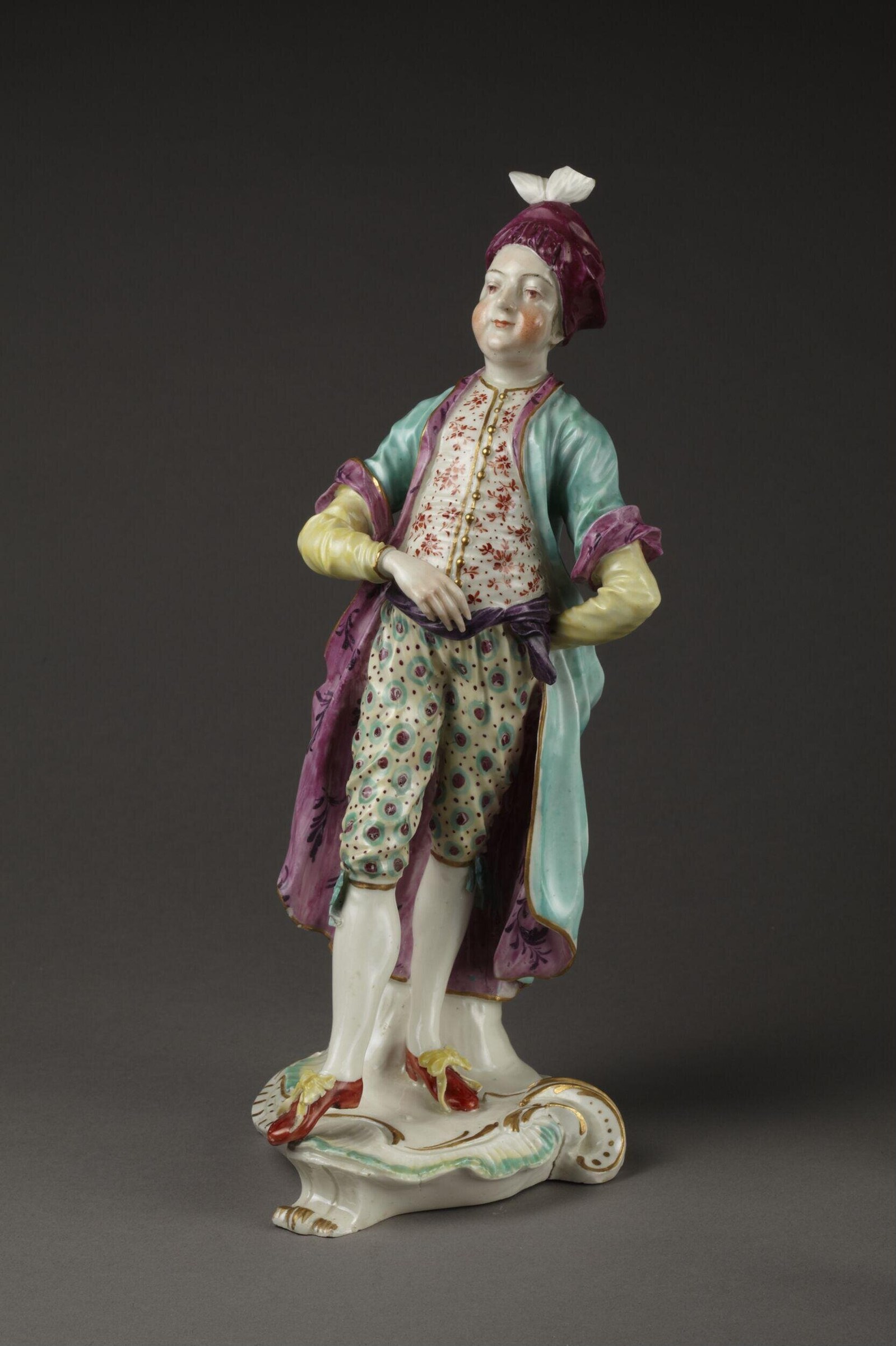 Porcelain figure of a man in 18th-century dress with patterned waistcoat, turquoise coat, and purple cap, standing in refined pose on a scrolled base.