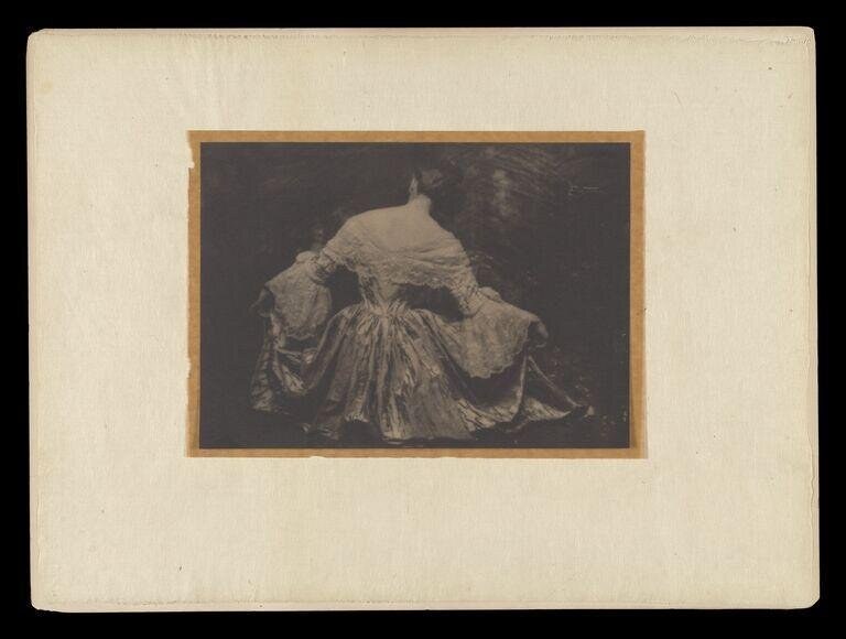 Woman in pale flowing gown stands alone, turned away, against dark background in dramatic light.