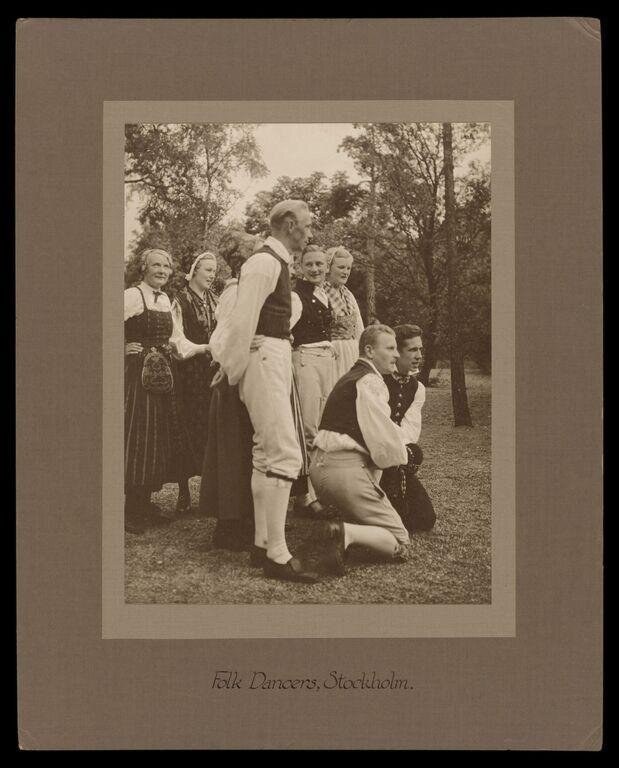 Seven people in Swedish folk costume stand and kneel in a woodland clearing, in a posed teaching arrangement.