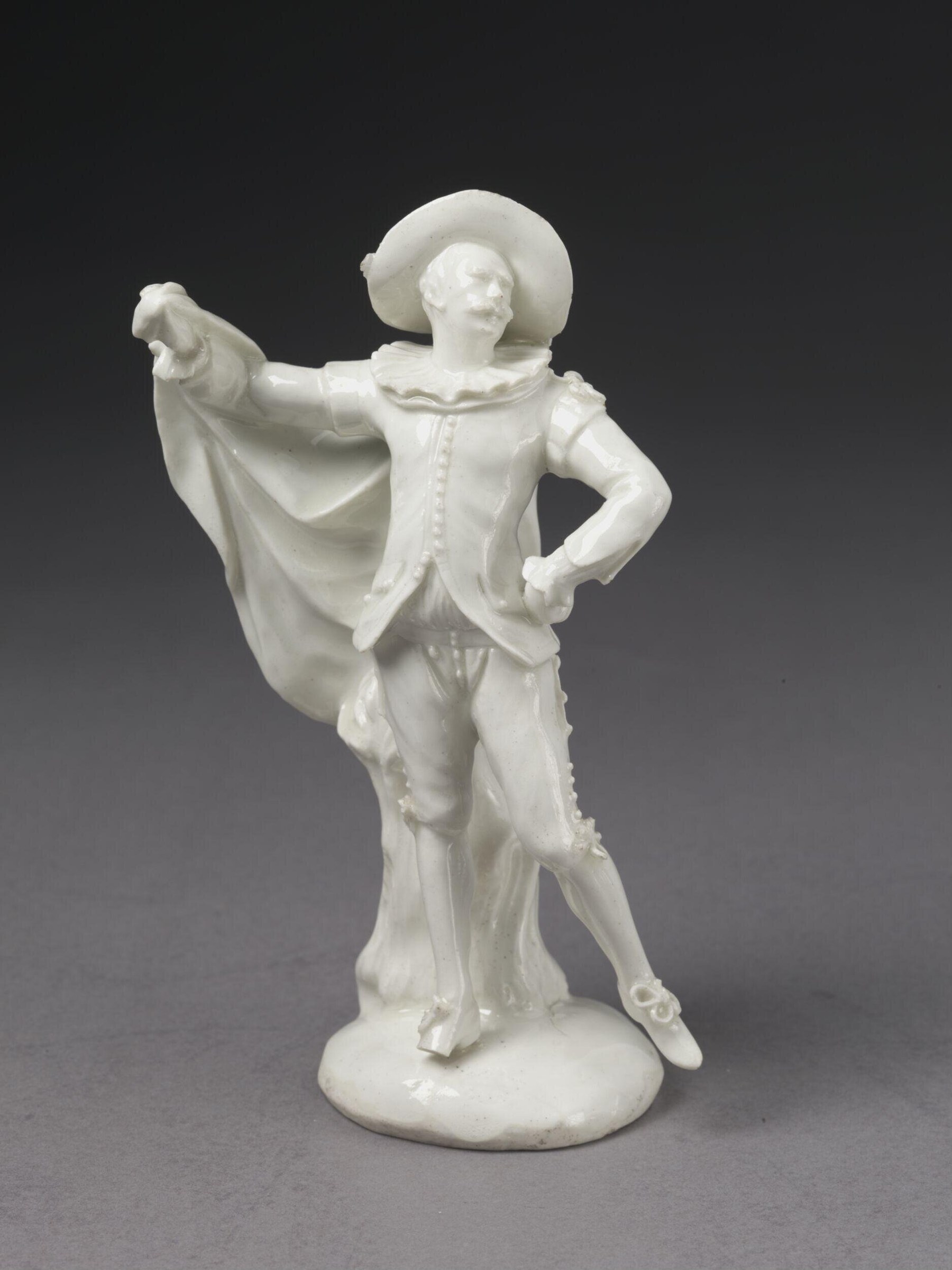 White porcelain figure of a masquerader in 18th-century costume, cape extended, hand on hip, in a theatrical display pose.