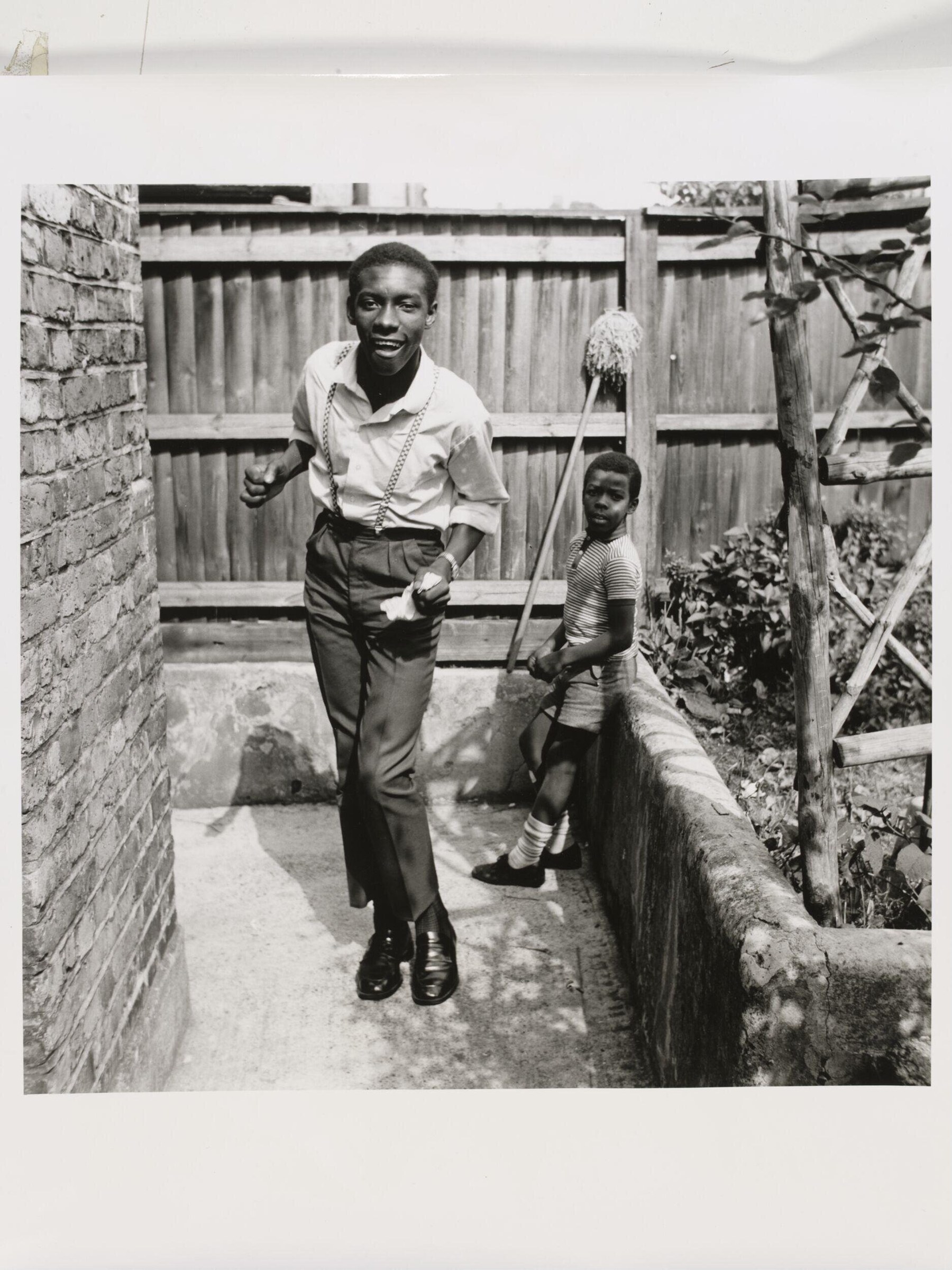 A teenage boy in a white shirt dances in a concrete yard; a younger boy watches from the side.
