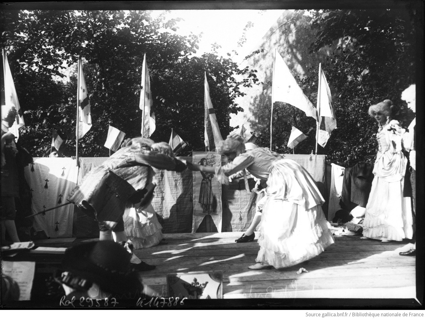 Two dancers in white leaning toward each other on a wooden platform surrounded by flags and spectators at an outdoor village celebration.