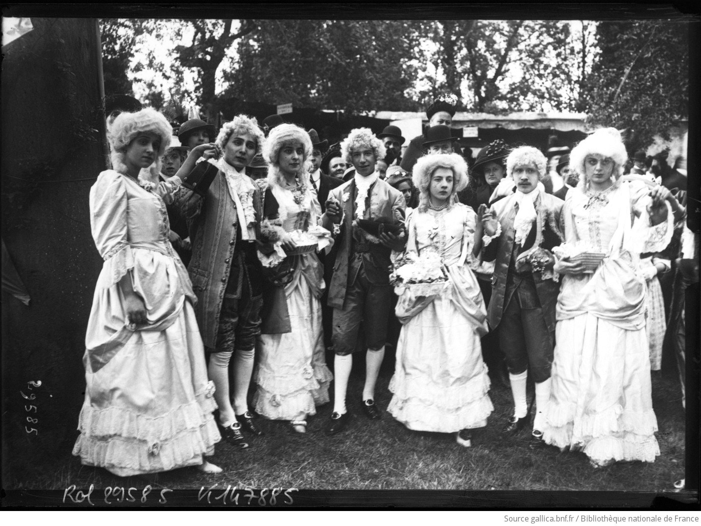 Eight costumed dancers in 18th-century dress—women in white gowns and men in dark coats, all wearing powdered wigs—stand in a formal line at an outdoor Breton