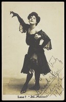 Male performer in drag costume with feathered hat, posing theatrically against plain background.