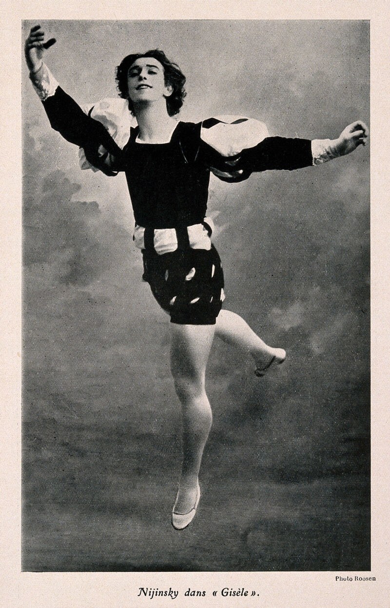 Male ballet dancer in mid-leap against gray background, arms spread wide, right leg extended high, head tilted back, wearing dark tunic and tights.