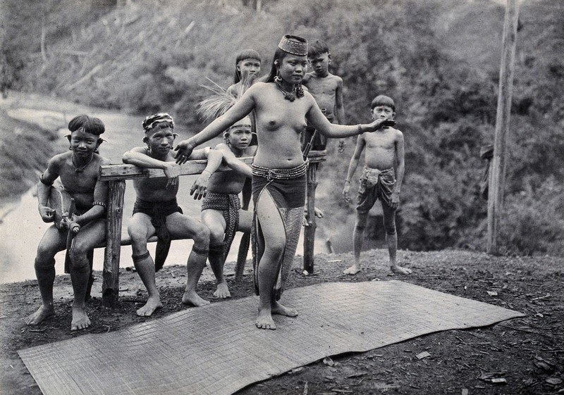 A bare-chested young woman with arms extended stands on a woven mat, surrounded by observing children in a tropical setting.