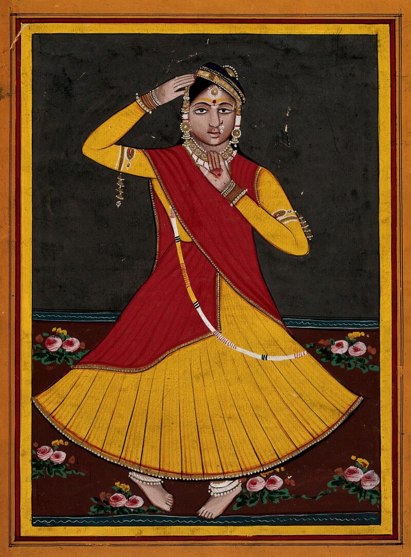 A woman in yellow pleated skirt, red shawl, and elaborate jewelry stands with her right arm raised, touching her decorative headpiece, against a dark