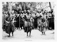 Black-and-white photograph of approximately a dozen African women in traditional dress gathered outdoors, with arms raised and bodies in varied postures