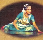 A female Bharatanatyam dancer in a teal silk blouse and pleated skirt sits in a composed pose, gazing downward, against a warm brown background.