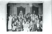 Formal group portrait of 18 people in 1930s dress posed indoors at a ballroom with ornate architectural details visible behind them.