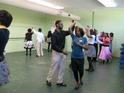 A mixed-age group of people in casual dress participate in a dance workshop in a bright studio, with a man and woman demonstrating in the center.